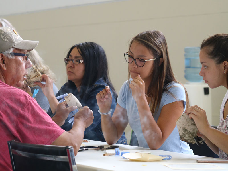 Young woman sitting at a table learning to make a birch bark basket from an elder