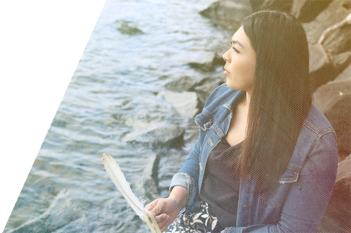 Anishinaabekwe holding an eagle feather, sitting on rocks next to the water