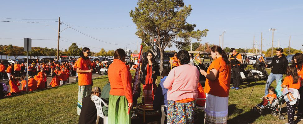 A group of women wearing ribbon skirts standing in a circle around a traditional drum