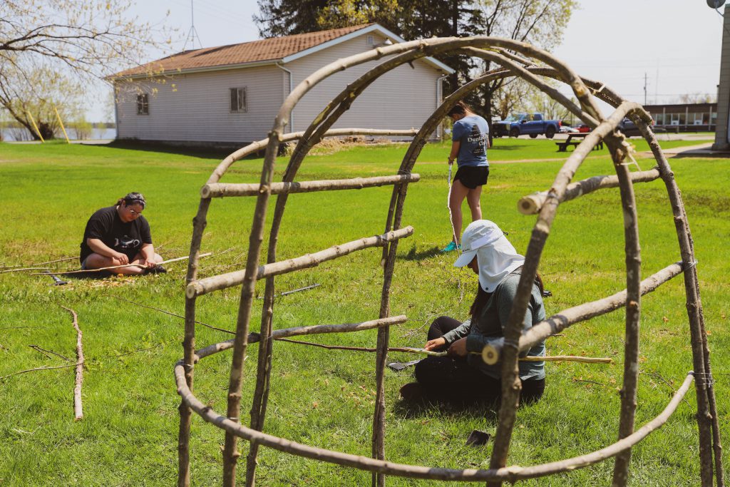 A group of individuals shaving sticks beside a traditional smoke hut structure