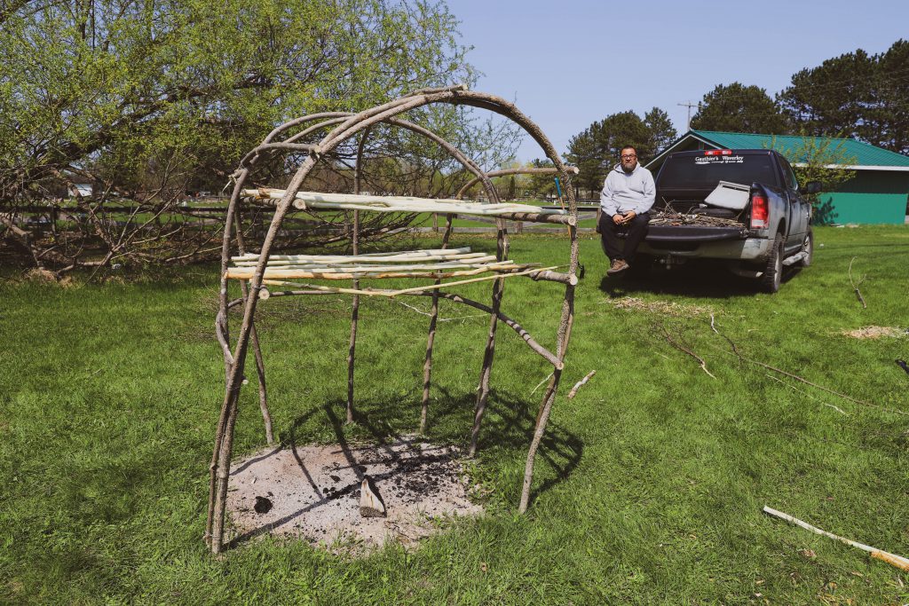 An Anishinaabe man sits behind a traditional smoke hut structure