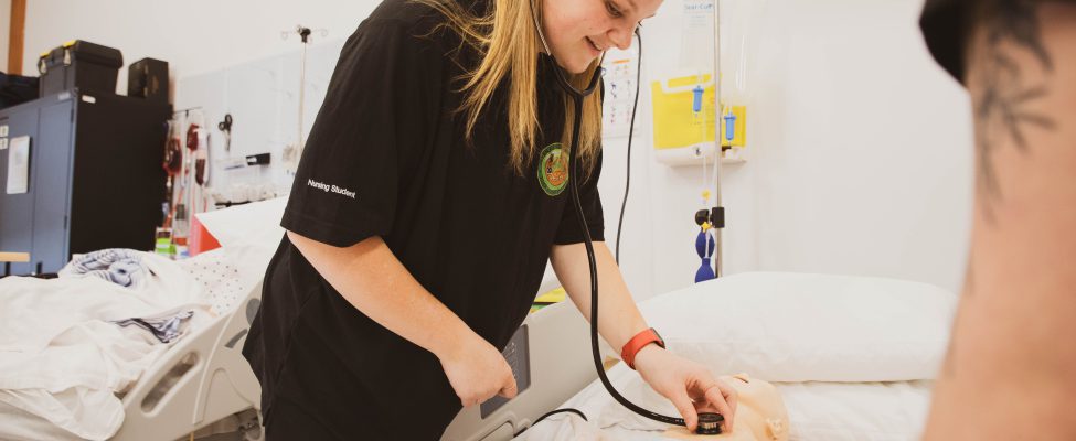 A practical nursing student uses a stethoscope during a sim lab.