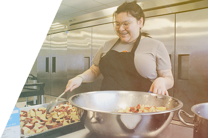 Woman smiling, working in a commercial kitchen holding a spoon stirring potatoes on a sheet tray.